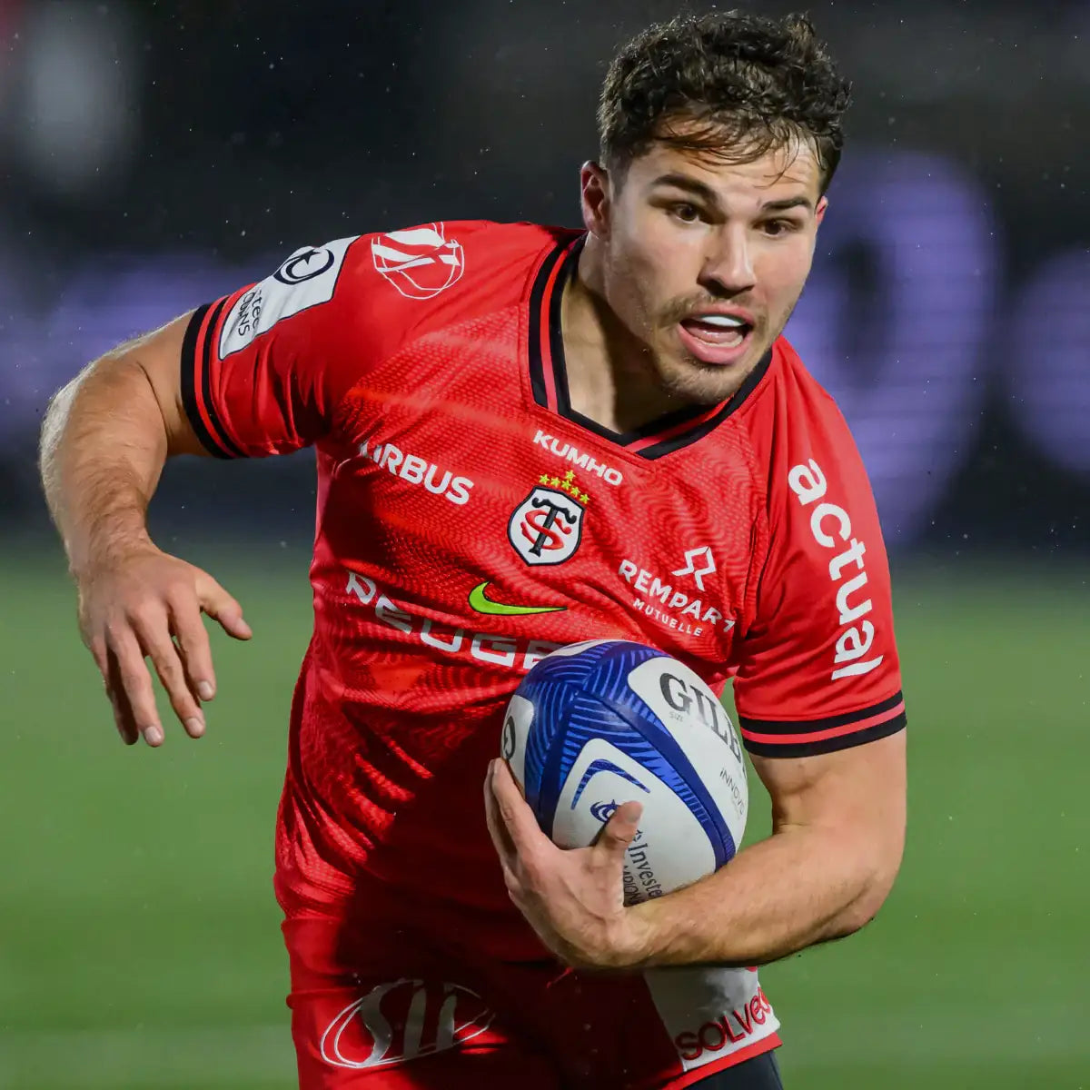 Rugby player in red jersey holding a ball on a blurred stadium background