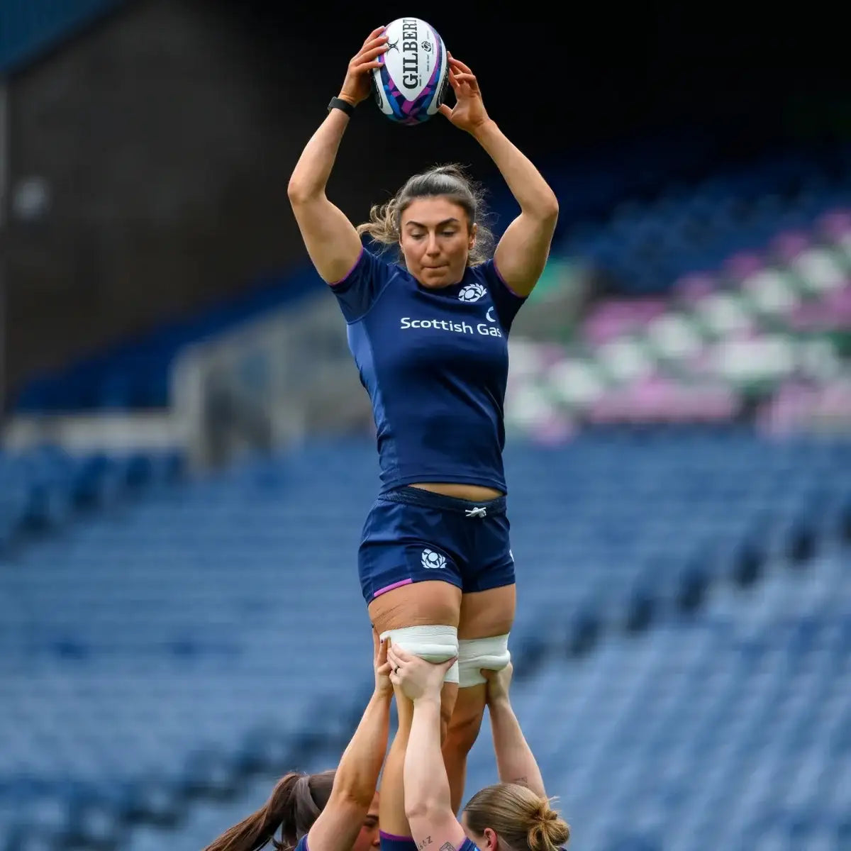 Woman in a blue sports uniform holding a rugby ball above her head in a stadium setting.