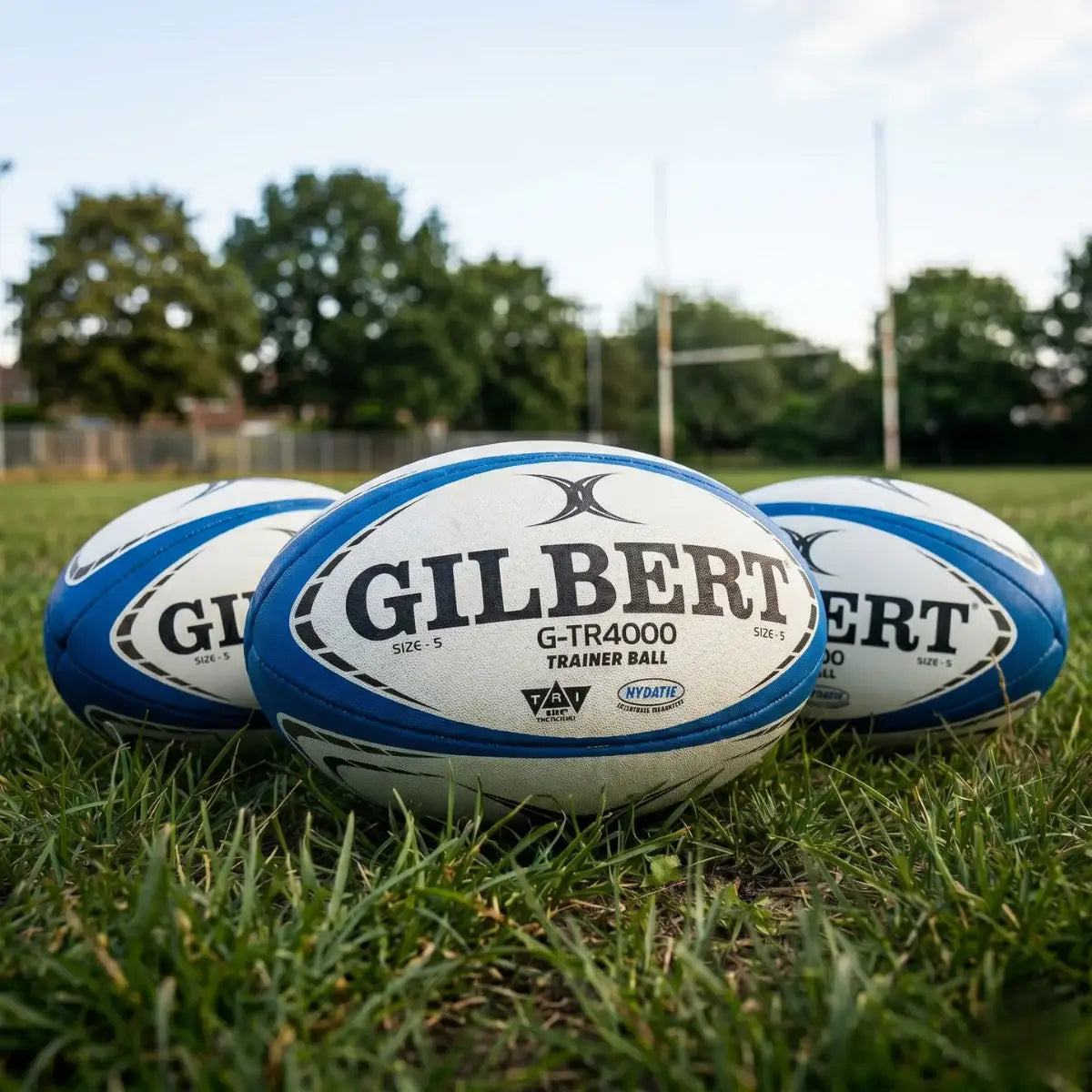Three Gilbert rugby balls on grass with a blurred sports field background