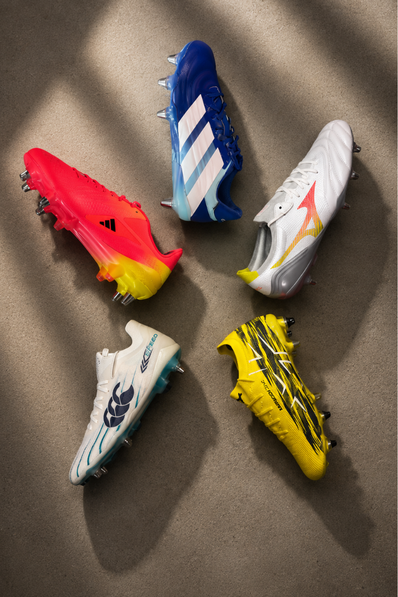 Five rugby boots in different colorways arranged on a textured gray surface with dramatic diagonal shadows. The shoes include red and yellow, blue and white, white and gray, white and teal, and yellow and black designs, each with visible brand logos.